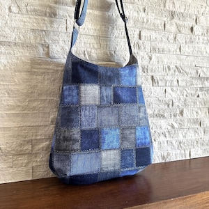 May include: A hobo-style shoulder bag constructed from a patchwork of various blue denim fabric squares. The bag features a long, adjustable black strap attached with silver-toned metal rings and visible stitching along the edges of each denim patch.