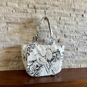 May include: A small tote bag made of off-white fabric featuring a black printed pattern of two birds perched on flowering branches. The design includes subtle script text and circular stamp motifs. It has two matching fabric handles at the top.