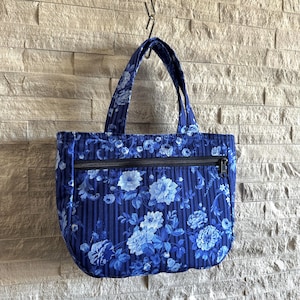 May include: A blue tote bag featuring a floral pattern with light blue and white flowers over a dark blue striped background. The bag includes two fabric handles and a horizontal black zipper pocket across the front exterior of the bag.