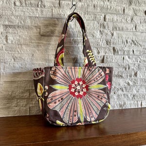 May include: A brown fabric tote bag featuring a large, stylized floral pattern in shades of pink, red, and light green. The bag has two matching fabric handles and a structured, rounded base, showcasing a repeating botanical print design.