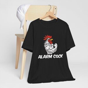 Puede incluir: Camiseta negra con un gallo musculoso, cresta roja y pico amarillo. El texto "ALARM COCK" está impreso en blanco. Un sombrero blanco descansa sobre una silla de madera en el fondo.
