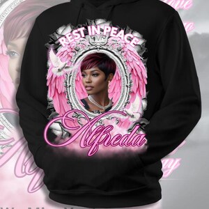 Pode incluir: Moletom preto com capuz com um retrato de uma mulher dentro de uma moldura com asas rosa, com as palavras "Rest in Peace" acima e "Alfreda" em it&aacute;lico abaixo. A frase "We Miss You!" est&aacute; na parte inferior.