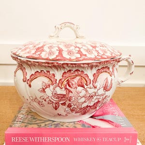 May include: A vintage ceramic chamber pot with a lid, featuring a pink and white floral transferware pattern and a curved handle. The pot rests on two stacked books, one titled "Reese Witherspoon Whiskey in a Teacup" and the other "Mastering the Art of French Cooking."