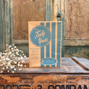 May include: A vintage paper package for "Truly Yours" brassieres, featuring a teal and cream color scheme. The package includes text that reads "styled for beauty, comfort, proper support." The package is set against a rustic wooden background.