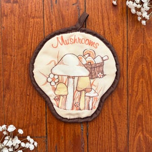 May include: A round fabric pot holder with a dark brown border and a hanging loop. The cream-colored center features a printed illustration of various mushrooms, small flowers, and a woven basket, with the word Mushrooms written in orange.