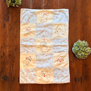 May include: A rectangular patchwork quilt featuring alternating squares of solid pale yellow fabric embroidered with line art of a dog, cat, mouse, elephant, clown, and teddy bear, and light blue fabric with a small floral pattern throughout.