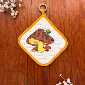 May include: A diamond-shaped quilted potholder featuring a white fabric with small orange dots and a central graphic of brown and yellow mushrooms with a small green insect. The potholder has a bright orange fabric border and a wooden hanging ring.