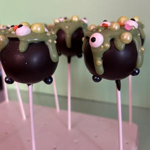 May include: Round black cake pops shaped like cauldrons on white sticks. Each features green icing dripping from the top, decorated with edible white candy eyeballs, small gold and pink spherical sprinkles, and tiny black feet.