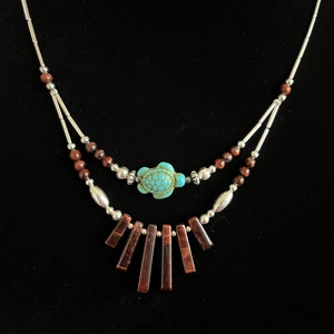 Puede incluir: Un collar de varias capas con un colgante de tortuga turquesa y cuentas rectangulares de color marrón rojizo. El collar tiene detalles plateados y pequeñas cuentas marrones, sobre un fondo negro. El diseño es simétrico y detallado.