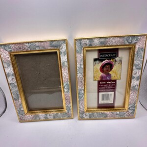 May include: Two rectangular photo frames with a floral pattern in shades of pink, green, and grey, featuring a gold-toned inner border. The right frame contains a display card with the text INTERCRAFT and a photo of a child in a pink hat.