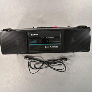 Könnte beinhalten: Ein schwarzer tragbarer Sanyo Radio-Kassettenrekorder mit zwei großen Lautsprechern an den Seiten, einem zentralen Bedienfeld mit dem Text SANYO, 2 WAY 4 SPEAKERS, AM/FM STEREO RADIO CASSETTE RECORDER und einem schwarzen Netzkabel mit Stecker.