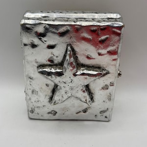 May include: A rectangular silver-colored metal lighter case featuring a prominent, raised five-pointed star centered on the front surface. The metal has a textured, hammered finish with irregular indentations and a metallic sheen throughout.