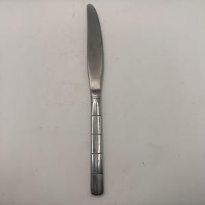 May include: A stainless steel dinner knife with a smooth, curved blade and a handle featuring a grid-like pattern of horizontal and vertical etched lines. The metal has a polished finish, and the design is consistent from the bolster to the base.
