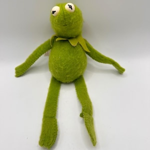 Puede incluir: Un peluche de rana de color verde brillante con un cuerpo de tela texturizada, brazos y piernas largos y delgados, y un cuello puntiagudo alrededor del cuello. La rana presenta dos grandes ojos blancos con pupilas negras y una boca cosida.