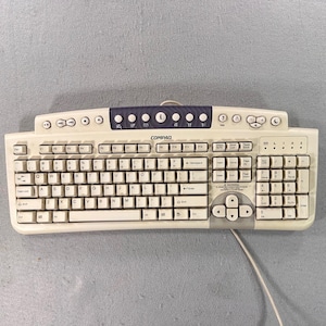May include: A beige Compaq computer keyboard with a QWERTY layout, a numeric keypad, and a dark blue top panel featuring various multimedia shortcut buttons, including an 'i' button, volume controls, and navigation keys with a wired connection.