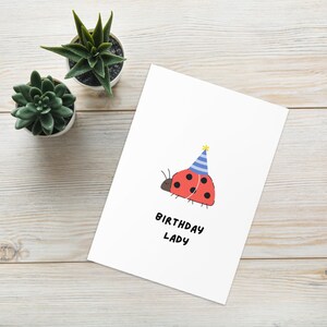 Könnte beinhalten: Eine weiße Grußkarte mit einer roten Marienkäfer-Illustration mit schwarzen Punkten, der einen blau-weiß gestreiften Partyhut mit gelbem Bommel trägt. Der Text "BIRTHDAY LADY" ist in schwarzen Großbuchstaben unter dem Marienkäfer gedruckt.