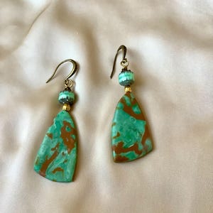 May include: A pair of dangle earrings featuring flat, triangular-shaped pendants with a marbled pattern of turquoise and brown. Each earring includes a small, ribbed turquoise bead above the pendant, connected by gold-toned hardware to a curved ear wire.