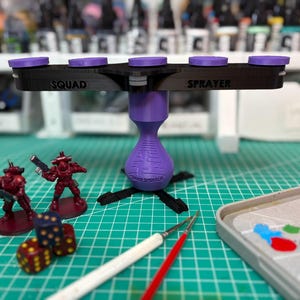 May include: A black and purple painting tool labeled SQUAD SPRAYER with five circular purple platforms on a horizontal bar supported by a purple base. Two red miniature figures, two multi-colored dice, and two paintbrushes are positioned nearby.