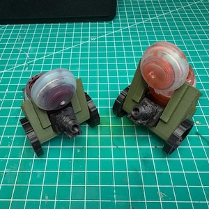 May include: Two miniature green and black toy cannons with clear and red-tinted tops, set against a green grid background. The cannons have a detailed design, suggesting a model or hobbyist item.