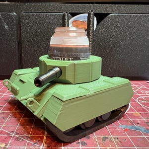 May include: A 3D-printed light green tank-shaped paint pot holder with a black barrel and two vertical black exhaust pipes. The turret features a circular cutout designed to securely hold a Citadel paint pot, which is currently inserted.