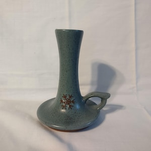 May include: A blue-grey ceramic candle holder with a speckled finish and a small handle on the side. The base features a brown snowflake-shaped design. The object has a wide, circular base that tapers upward into a narrow, cylindrical neck.