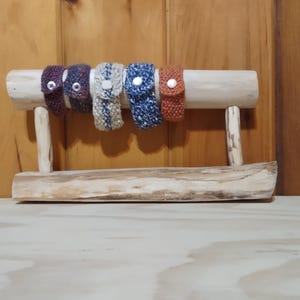 May include: A wooden display stand holds six knitted wristbands in various colors: purple, gray, white, blue, and orange. Each wristband has a button closure. The stand is made of natural wood and sits on a wooden surface.