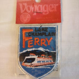 May include: A vintage Lake Champlain Ferry patch with a shield shape. The patch features a blue background with an orange "F" and the words "Lake Champlain Ferry" in black. A ferry boat is depicted in white and orange. The patch is attached to a red Voyager card.