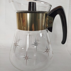 May include: Vintage glass coffee carafe with a clear body, gold-toned metal band, and black handle. The carafe features a starburst pattern and a clear glass lid. A classic mid-century modern design.