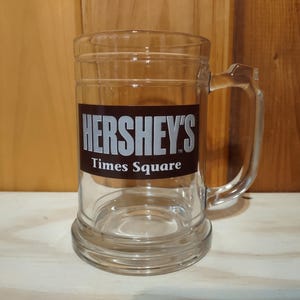 May include: Clear glass beer mug with a handle. The mug features the Hershey's Times Square logo in white lettering on a brown rectangular label. The mug is empty and sits on a light-colored surface.