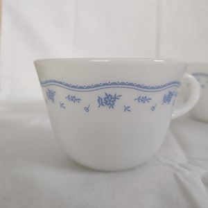 May include: White ceramic teacup with a blue floral pattern and a wavy blue border. The cup has a handle and is set against a white background. A second cup is partially visible in the background.