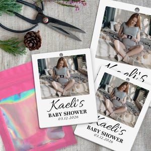 May include: White polaroid-style baby shower invitations with a photo of a pregnant person holding an ultrasound image. The invitations are labeled "Kaelis BABY SHOWER 03.12.2026". A pink iridescent pouch and scissors are also in the image.