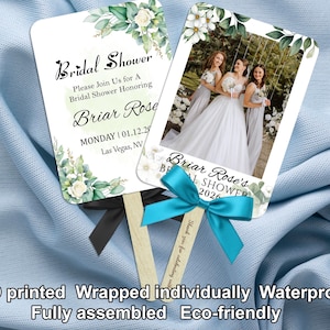 May include: Two white bridal shower fans with floral and greenery designs. One fan displays a photo of a bride and bridesmaids. Text includes "Bridal Shower" and "Briar Rose". The fans are attached to wooden sticks with ribbons.