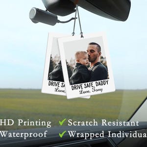 May include: Two rectangular car air fresheners hanging from a rearview mirror. Each features a photo of a man and child with the text "DRIVE SAFE, DADDY Love, Tony". The fresheners are white with black text and a photo.