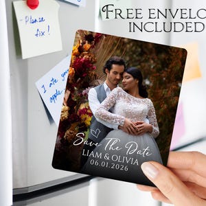 May include: A square magnet featuring a couple embracing, with the text "Save The Date" and the names "Liam & Olivia" along with the date "06.01.2026". The magnet is held up against a refrigerator. The text "Free envelopes included" is also visible.