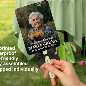 Puede incluir: Un abanico conmemorativo con una fotograf&iacute;a de una mujer sonriendo y sosteniendo flores, con el texto "In Loving Memory of MARIA GREEN 11.02.1954-05.27.2024." El abanico tiene una cinta negra y un mango de madera.
