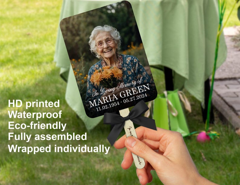 Puede incluir: Un abanico conmemorativo con una fotograf&iacute;a de una mujer sonriendo y sosteniendo flores, con el texto "In Loving Memory of MARIA GREEN 11.02.1954-05.27.2024." El abanico tiene una cinta negra y un mango de madera.