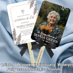 Puede incluir: Dos abanicos conmemorativos con mangos de madera y lazos negros. Un abanico presenta una foto de una anciana sosteniendo flores, con el texto "In Loving Memory of Maria Green". El otro abanico tiene el texto "Please join us for a Celebration of Life."