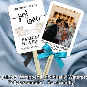 Puede incluir: Dos abanicos de boda blancos con mangos de madera. Un abanico tiene el texto "NOTHING FANCY just love" y los nombres Samuel Heath y la fecha de la boda. El otro abanico tiene una foto de una pareja besándose. Ambos abanicos tienen una cinta.