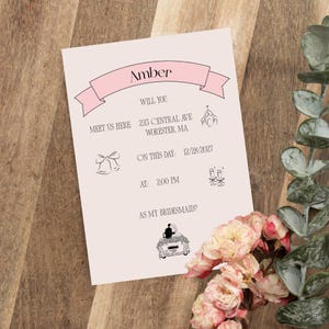以下が含まれることがあります： 「Amber」という名前が書かれた、薄いピンク色の招待状。招待状には「ここで私たちに会いませんか」というテキストと、日付、時間、場所など、結婚式の詳細が記載されています。花と緑のアクセントが招待状を飾っています。