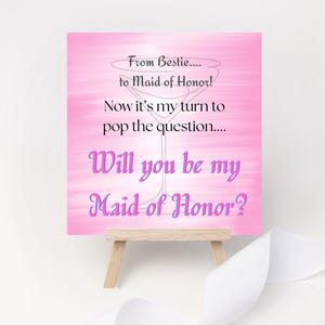 May include: A pink card on a small wooden easel with the text "From Bestie... to Maid of Honor! Now it's my turn to pop the question... Will you be my Maid of Honor?" A white ribbon is in the background.