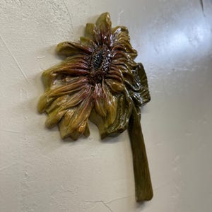 May include: A detailed, three-dimensional sunflower sculpture with a textured surface. The petals transition from yellow-green to reddish-brown, with a dark centre. The stem is a muted green, and the artwork is mounted on a white wall.
