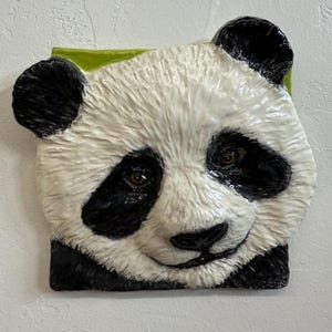 May include: A ceramic panda bear face sculpture with black and white fur, black ears and eye patches, and brown eyes. The panda is set against a green background.
