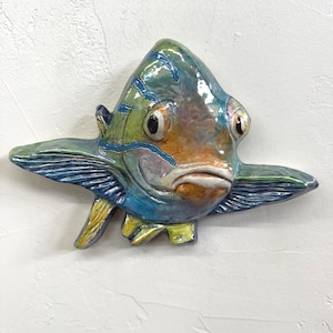 Op de afbeelding: Een keramische vissenbeeld met een kleurrijk ontwerp. De vis heeft tinten blauw, groen, geel en oranje. De sculptuur heeft grote ogen en vinnen en is op een witte muur gemonteerd.