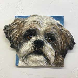 以下が含まれることがあります： 茶色と白の毛並みのシーズー犬のセラミック製の壁飾り。犬の目は茶色で鼻は黒です。彫刻は青い背景に設置されています。