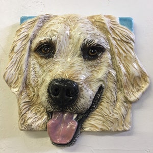 Può includere: Una scultura da parete in ceramica della testa di un golden retriever. Il cane ha un manto bianco e crema, un naso nero e una lingua rosa che sporge. La scultura è montata su uno sfondo blu.