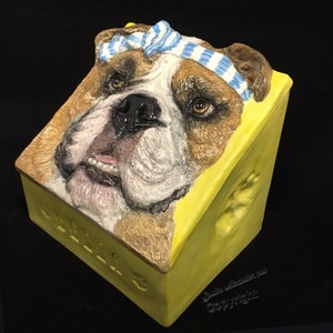 Op de afbeelding: Een gele keramische doos met een bulldoggezicht aan de voorkant. De bulldog draagt een blauw-wit gestreepte strik. De doos heeft het woord "Cadeaus" aan de voorkant en een pootprint aan de zijkant. De doos is gemaakt door Sandra Alexander.