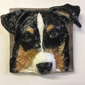 Può includere: Una scultura in ceramica di una testa di cane. Il cane ha il pelo nero, bianco e marrone. La scultura è dettagliata e realistica.
