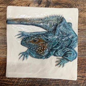 May include: A ceramic square tile featuring a detailed, three-dimensional depiction of a blue iguana. The iguana has a textured, scaly appearance with brown and blue coloration. The tile is white with a glossy finish, and the iguana is positioned diagonally.