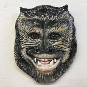 May include: A ceramic wall hanging in the shape of a monkey face. The monkey has brown eyes, a wide grin with visible teeth, and fur-like texture in shades of gray and beige. The piece has a glossy finish.