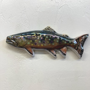 May include: A ceramic fish sculpture with a glossy finish. The fish has a speckled pattern in shades of yellow, green, and red, with a dark blue-green back and orange fins. The sculpture is mounted on a white textured surface.